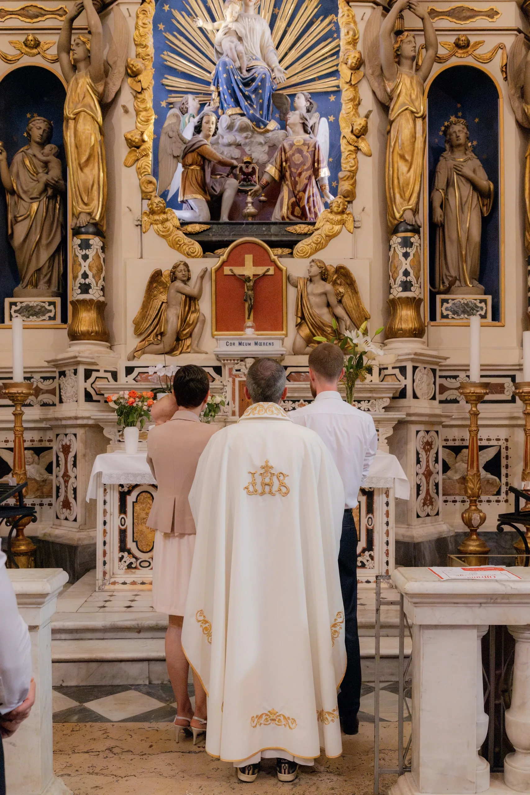 Vus de dos devant le retable doré, le prêtre en chasuble blanche, une femme en veste beige et un homme en chemise blanche