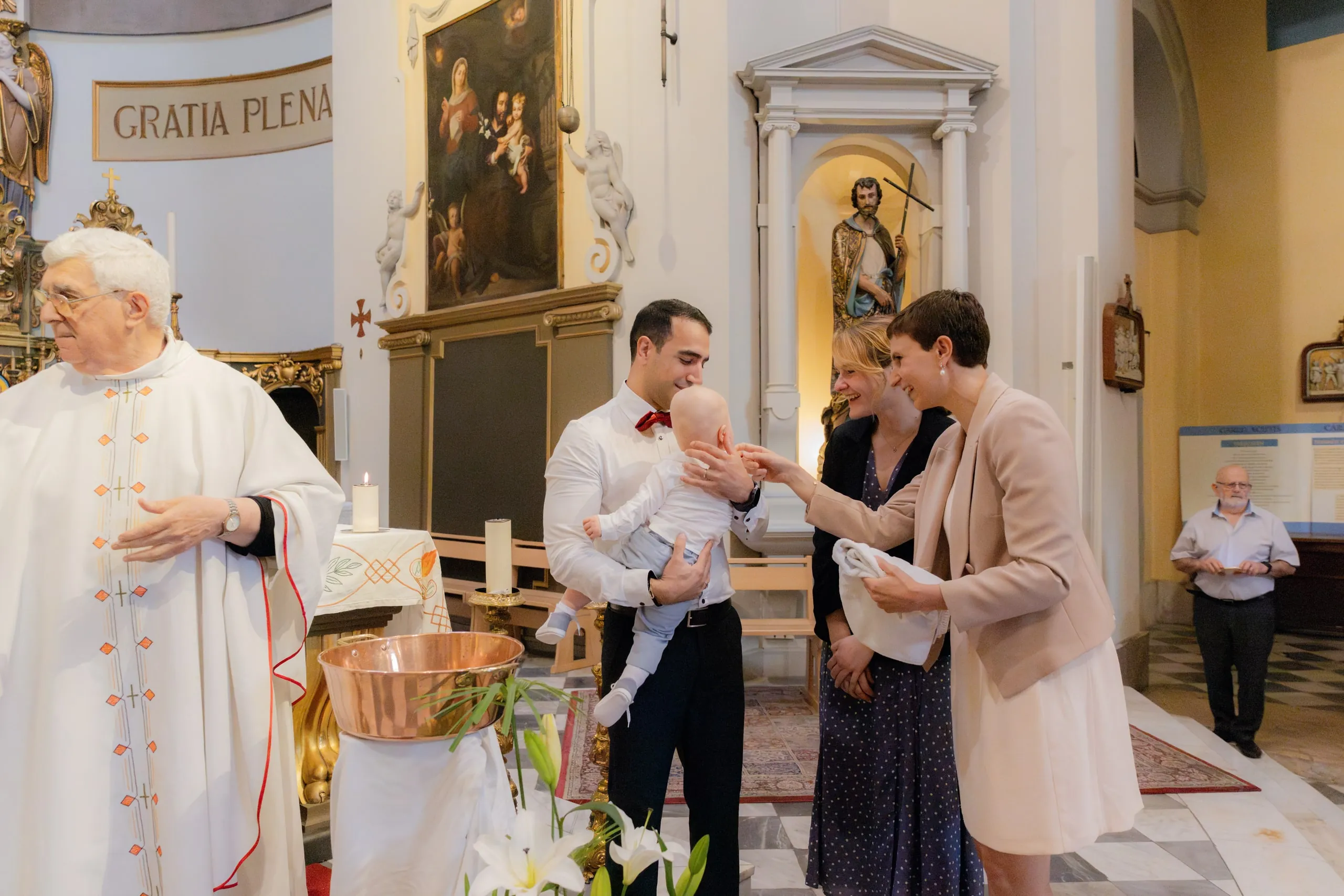 Le prêtre en chasuble blanche et, face à lui, un père au nœud papillon rouge avec son bébé, entouré de deux femmes