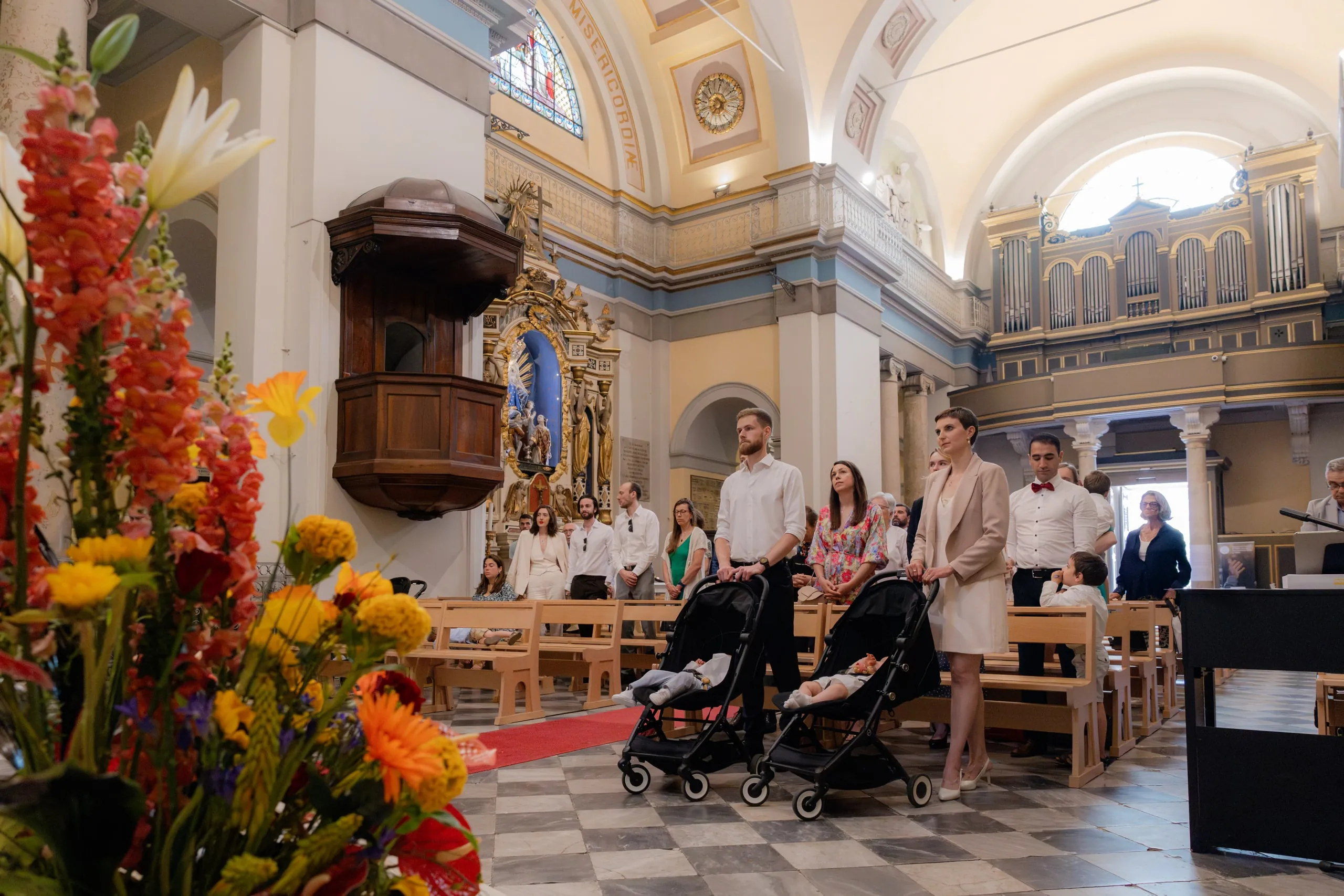 La nef de l'église derrière un bouquet de fleurs orangées, les invités debout et deux poussettes au fond