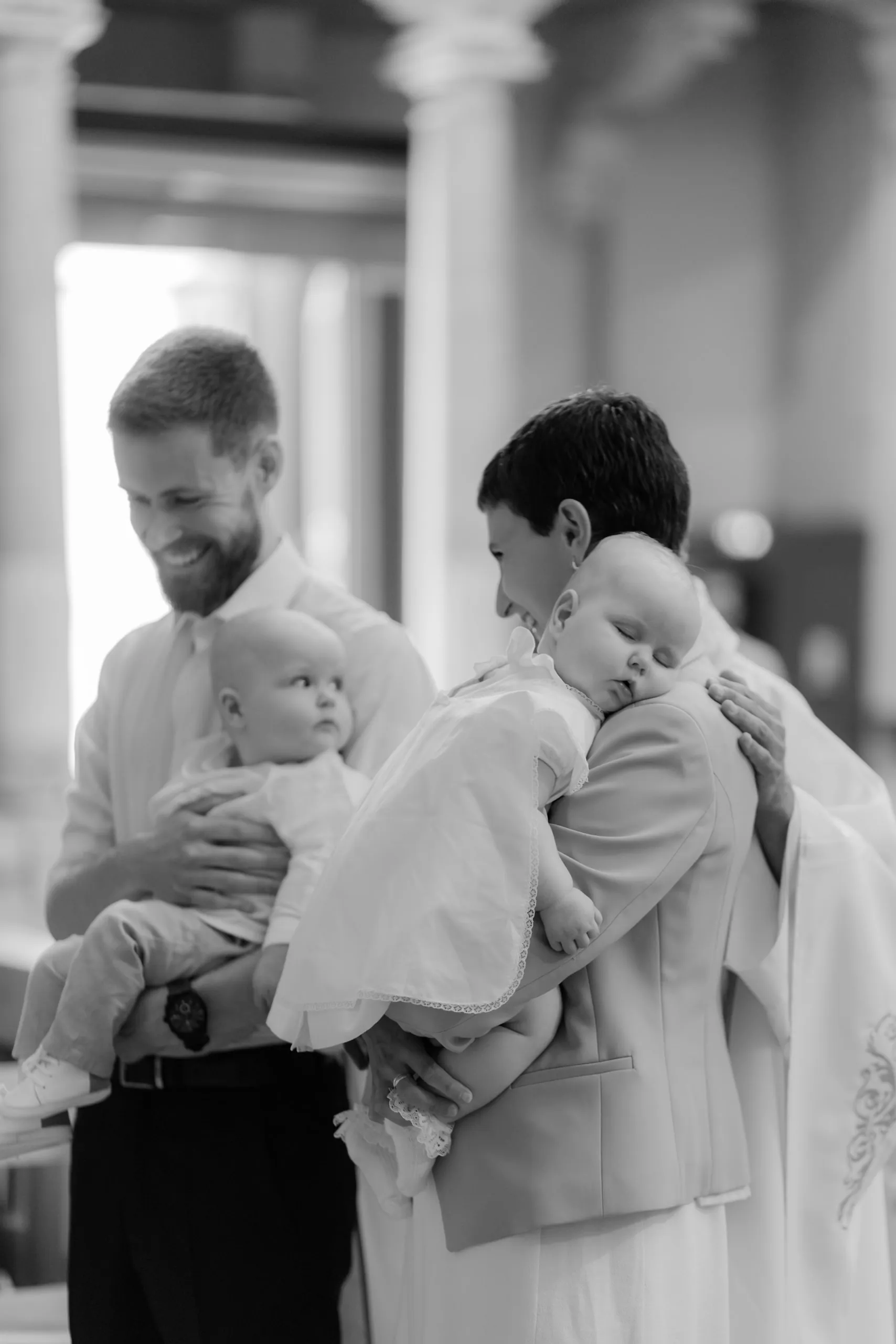 En noir et blanc, un homme barbu sourit à son bébé tandis qu'une femme serre contre elle un bébé endormi en robe de baptême