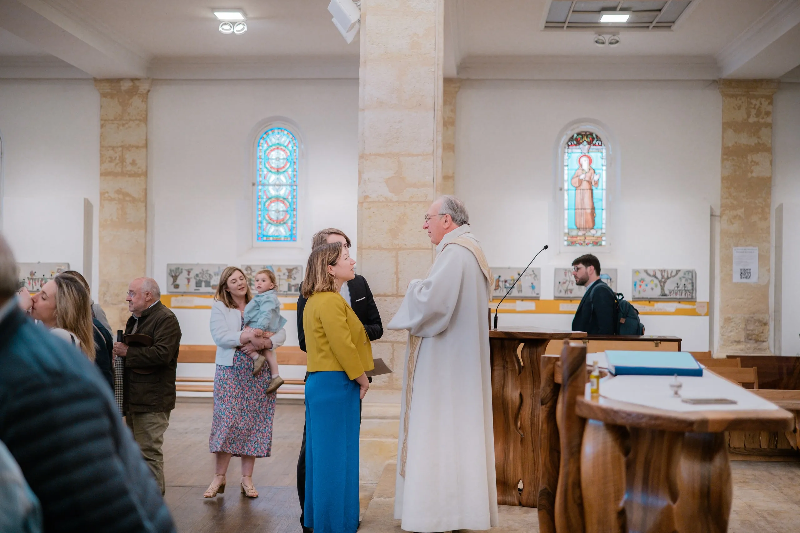 Sous les vitraux de la chapelle, le prêtre s'entretient avec une femme en veste jaune, pendant que les invités discutent