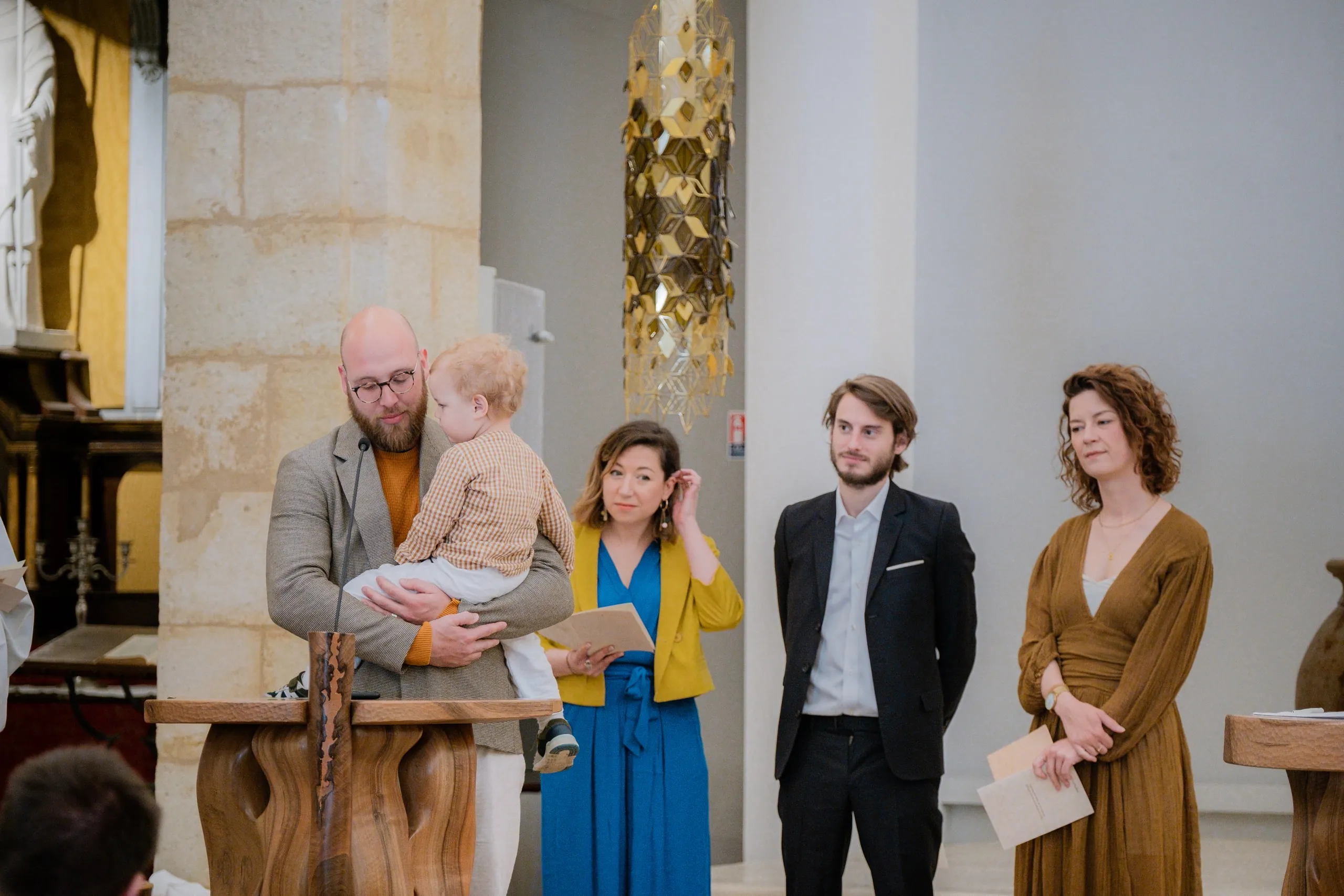 Dans la chapelle, le père portant son petit garçon au pupitre, une femme en veste jaune, un homme en costume sombre et la mère