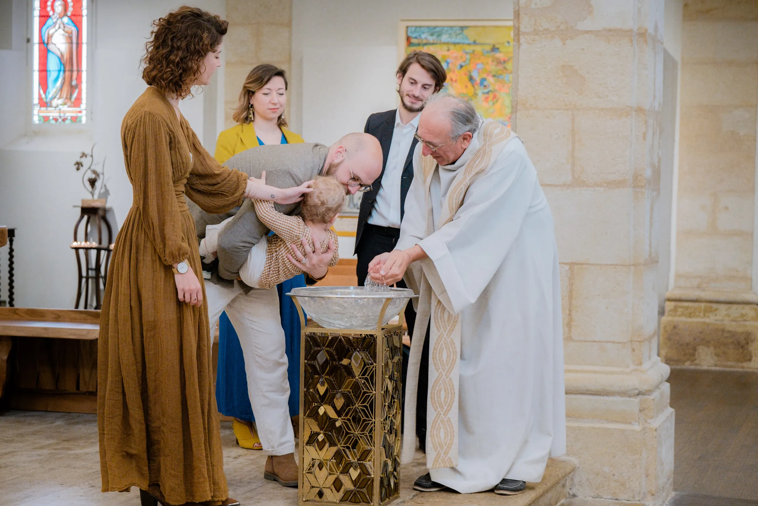 Le petit garçon, penché par son père au-dessus de la vasque, reçoit l'eau du baptême versée par le prêtre, sous le regard de sa mère